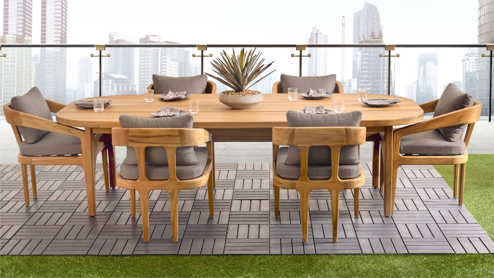 An oval-shaped wooden dining table with eight light-brown teakwood chairs is set on a gray-toned, interlocking, decking-style flooring. 