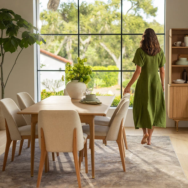 A woman in a green dress stands by a large window in a dining room with a wooden table, cream chairs, and a potted plant, looking outside.