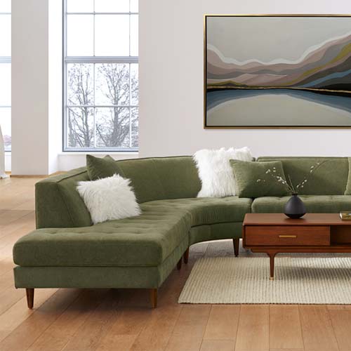 A green sectional sofa with white pillows and a wooden coffee table sit on a rug in a bright living room with large windows and a landscape painting on the wall.